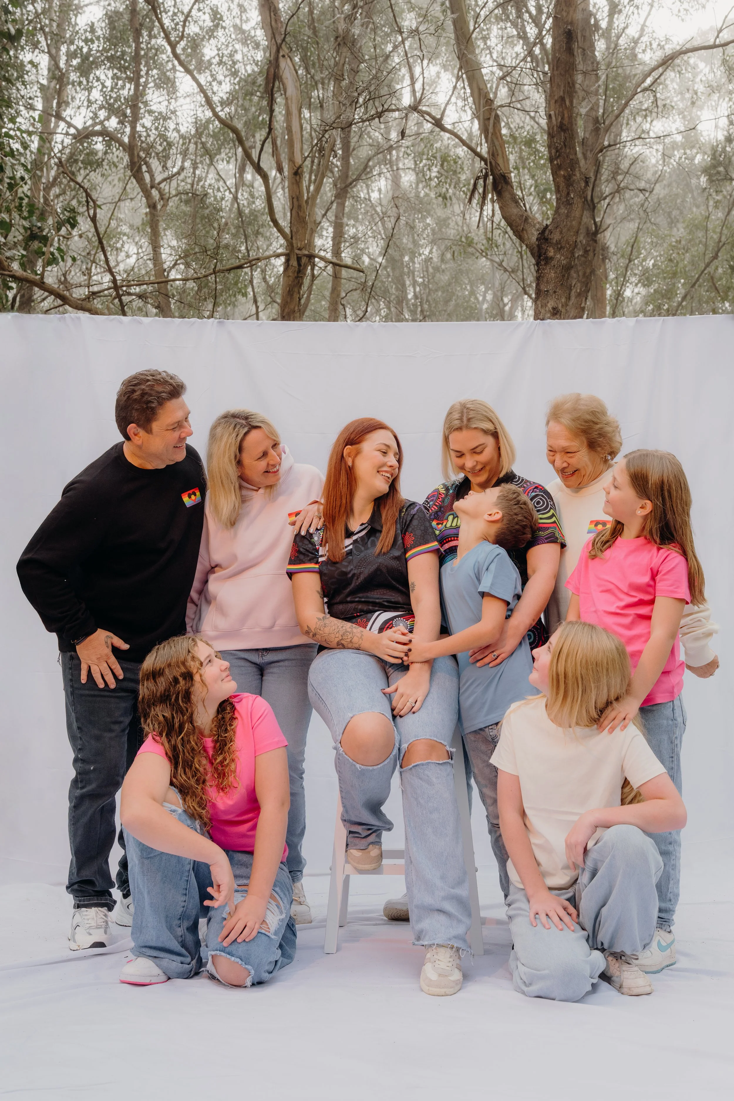 Family photo outdoors with a group of adults and children smiling and laughing together, against a white backdrop with trees in the background.