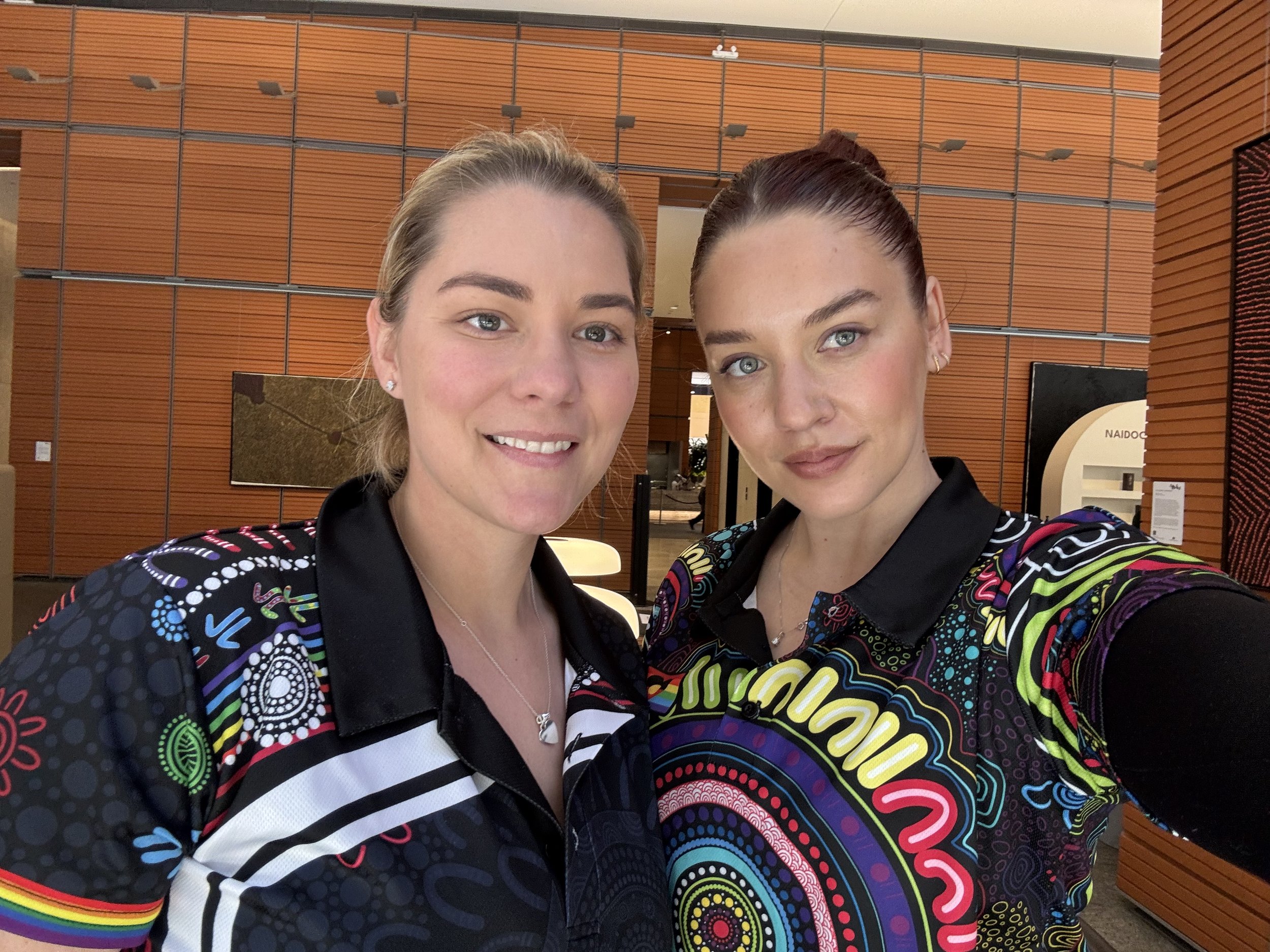 Two women with braided hair and colorful shirts smiling at the camera in a modern indoor setting with wood-paneled walls.