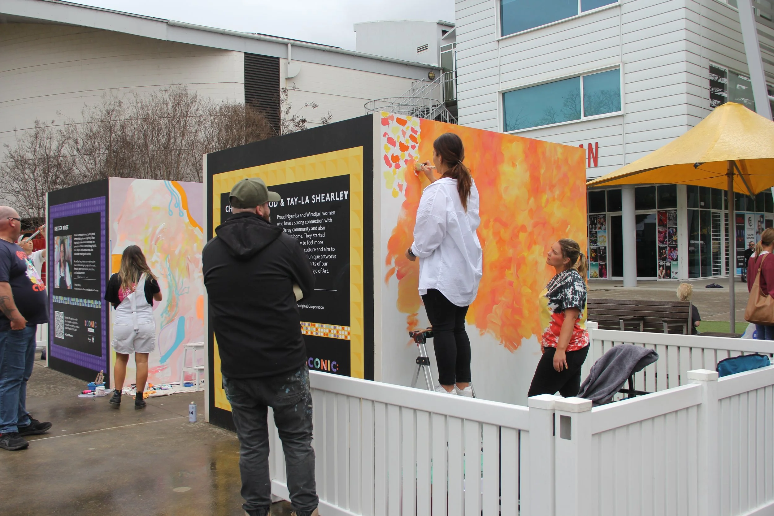 People painting murals on large display boards at an outdoor event, with some observing and others actively working on artwork.