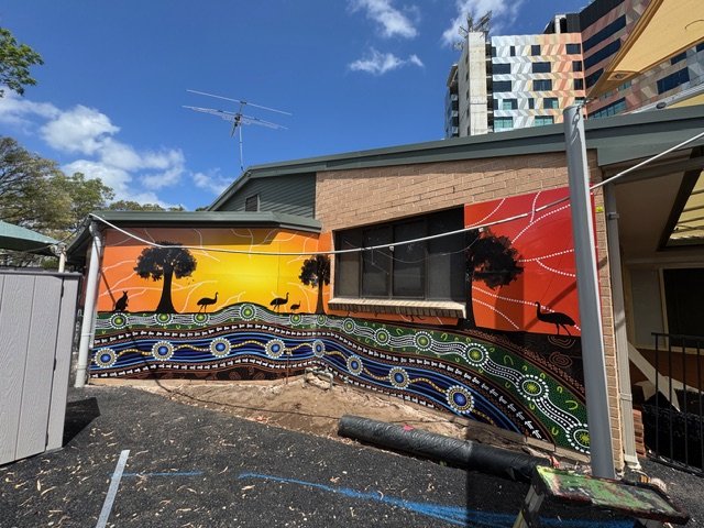 Mural with abstract pattern and sunset scene of trees and animals on exterior wall of building.