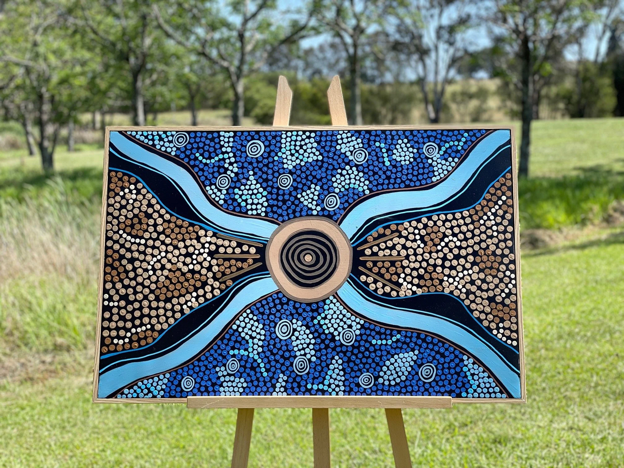 A painting with Aboriginal dot art styles on an easel outdoors, featuring a central circular pattern and symmetrical lines extending outward in blue, black, white, and brown colors, with a background of grassy area and trees.