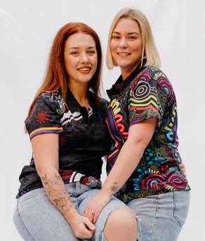Two young women with different hair colors, wearing colorful patterned shirts and light jeans, posing together against a plain white background.