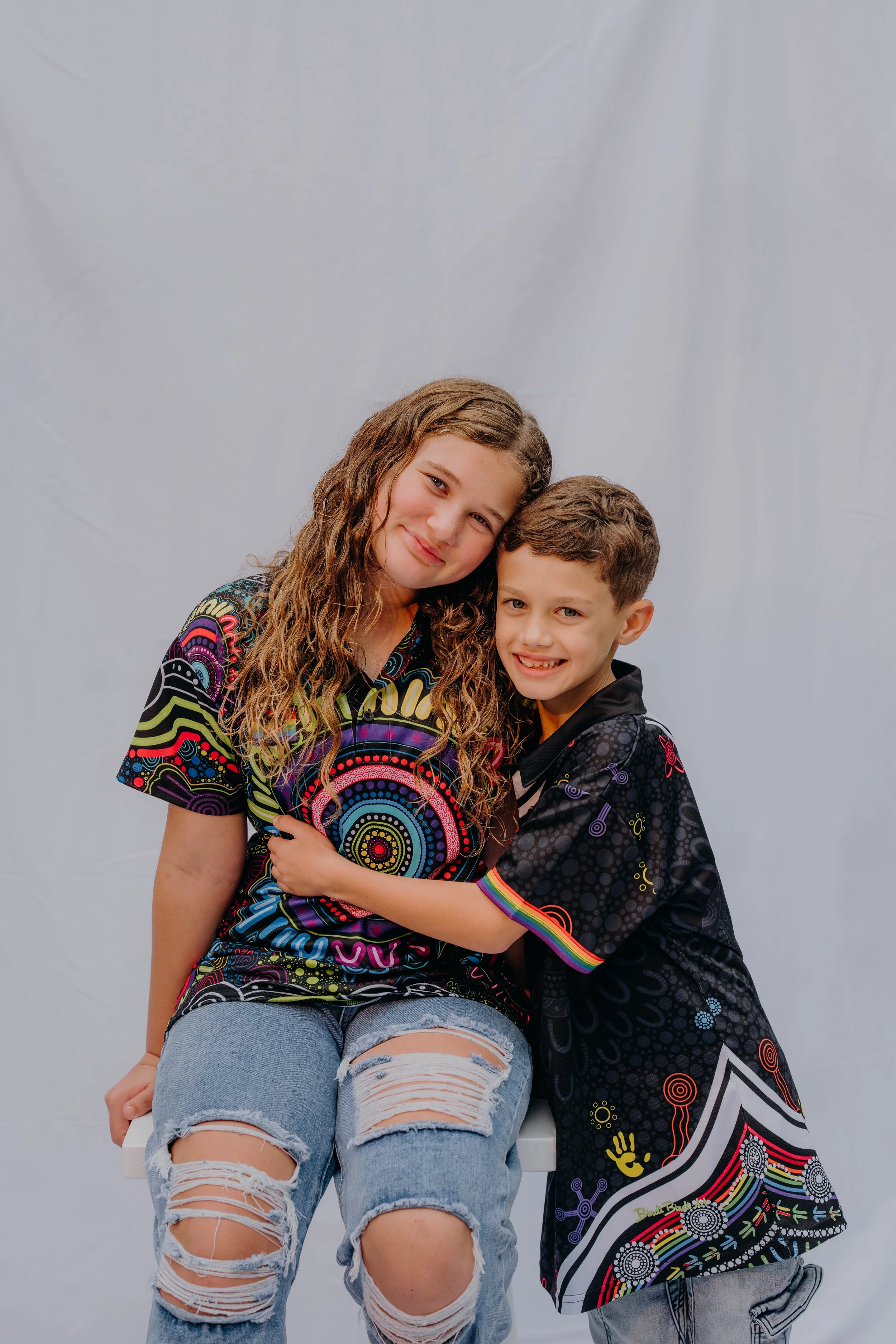A girl and a boy hugging and smiling at the camera against a plain light background. The girl is wearing a colorful, patterned shirt and ripped jeans, while the boy is dressed in a black shirt with colorful designs.