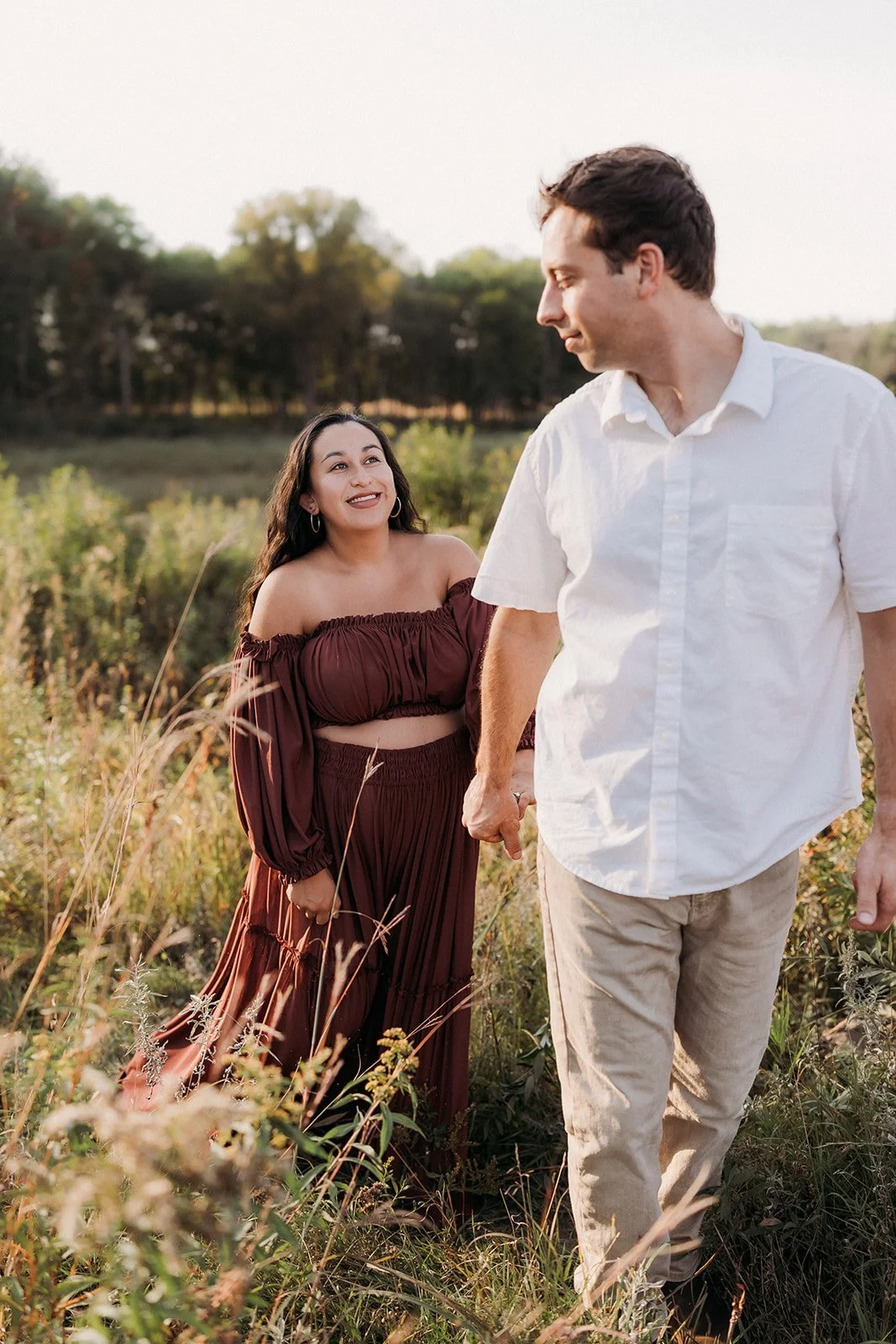 A couple holding hands in a field with greenery, the woman is wearing a brown off-the-shoulder outfit, and the man is dressed in a white shirt and beige pants, with a sunset sky in the background.