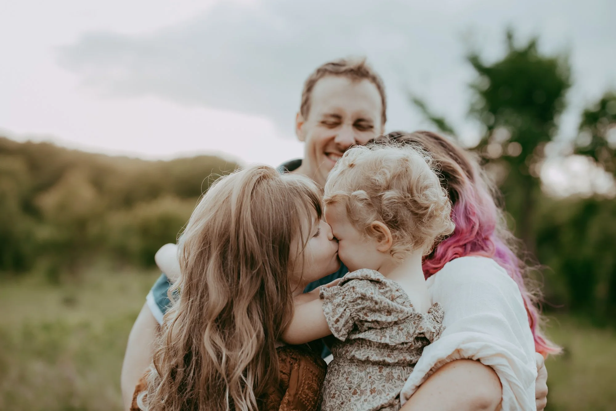 two sisters kiss each other while being held by their mom and dad at Crow Hassan Park in Hanover, MN during a family photo shoot with Ann Marie of AMG Photography