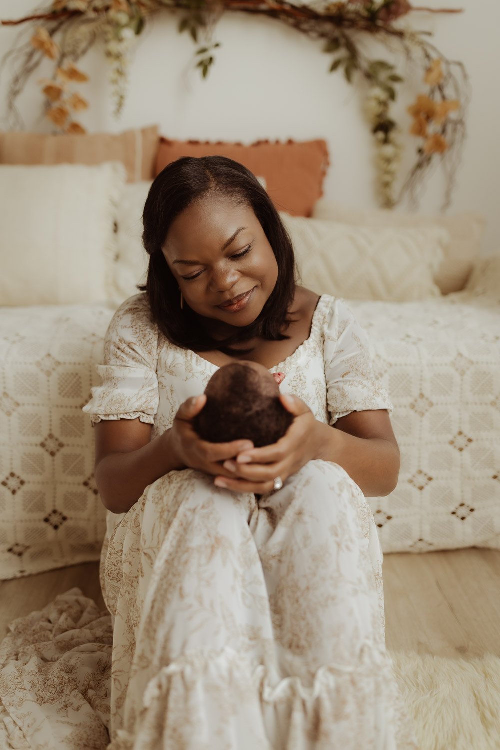 A mother sits in front of a bed decorated with pink and lace pillows and holds her baby girl on her knees.