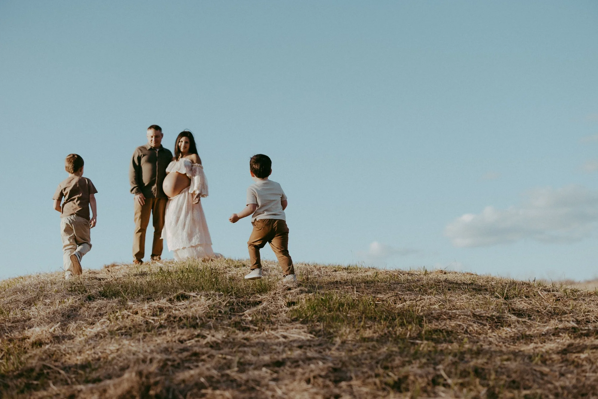 A pregnant woman standing outdoors with three young boys around her on a grassy hill under a clear blue sky.