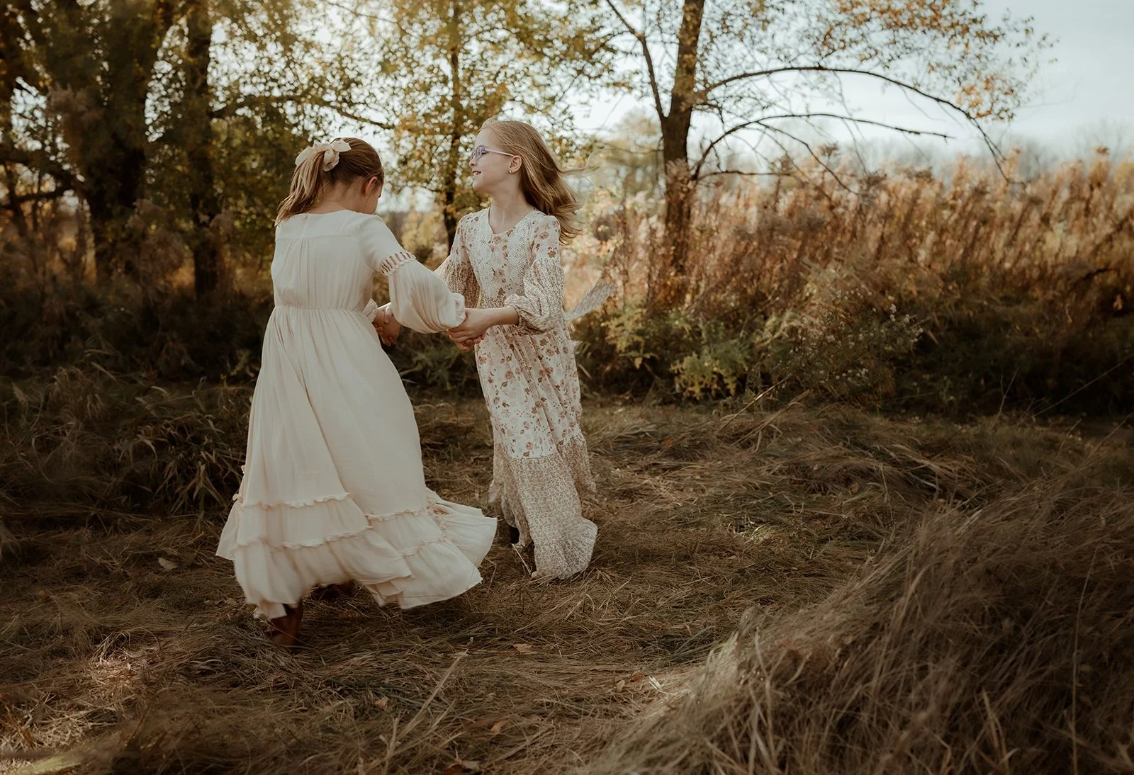 Two women in vintage dresses holding hands and dancing outdoors in a fall forest.