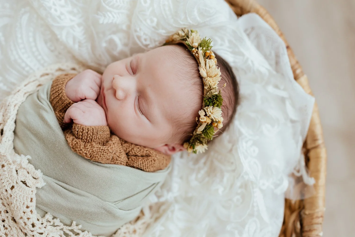 a baby girl in a light green and cream swaddle is wearing a flower crown and sleeping in a basket