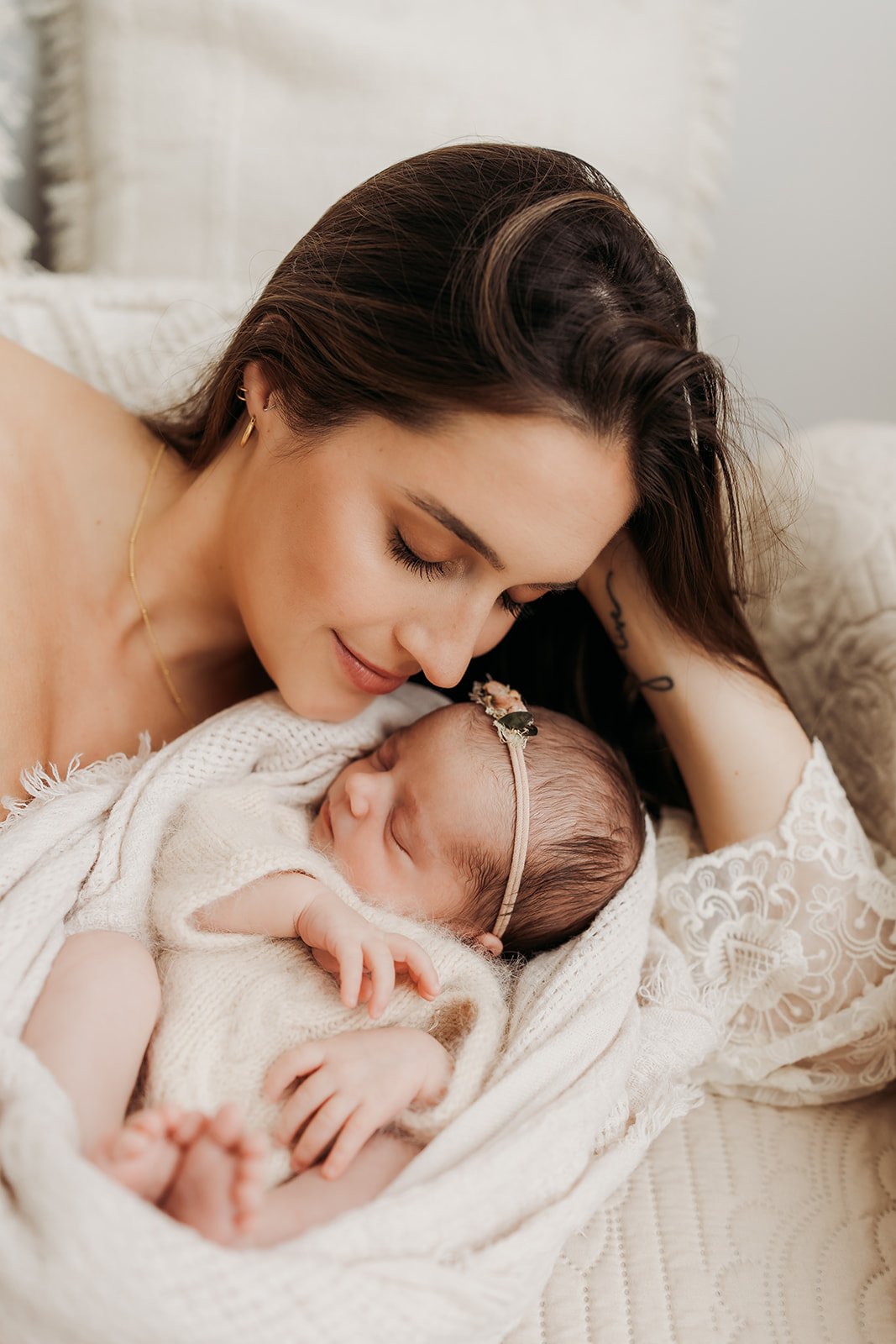  A mother in a white lace dress snuggles her newborn baby girl, who is sleeping next to her. 