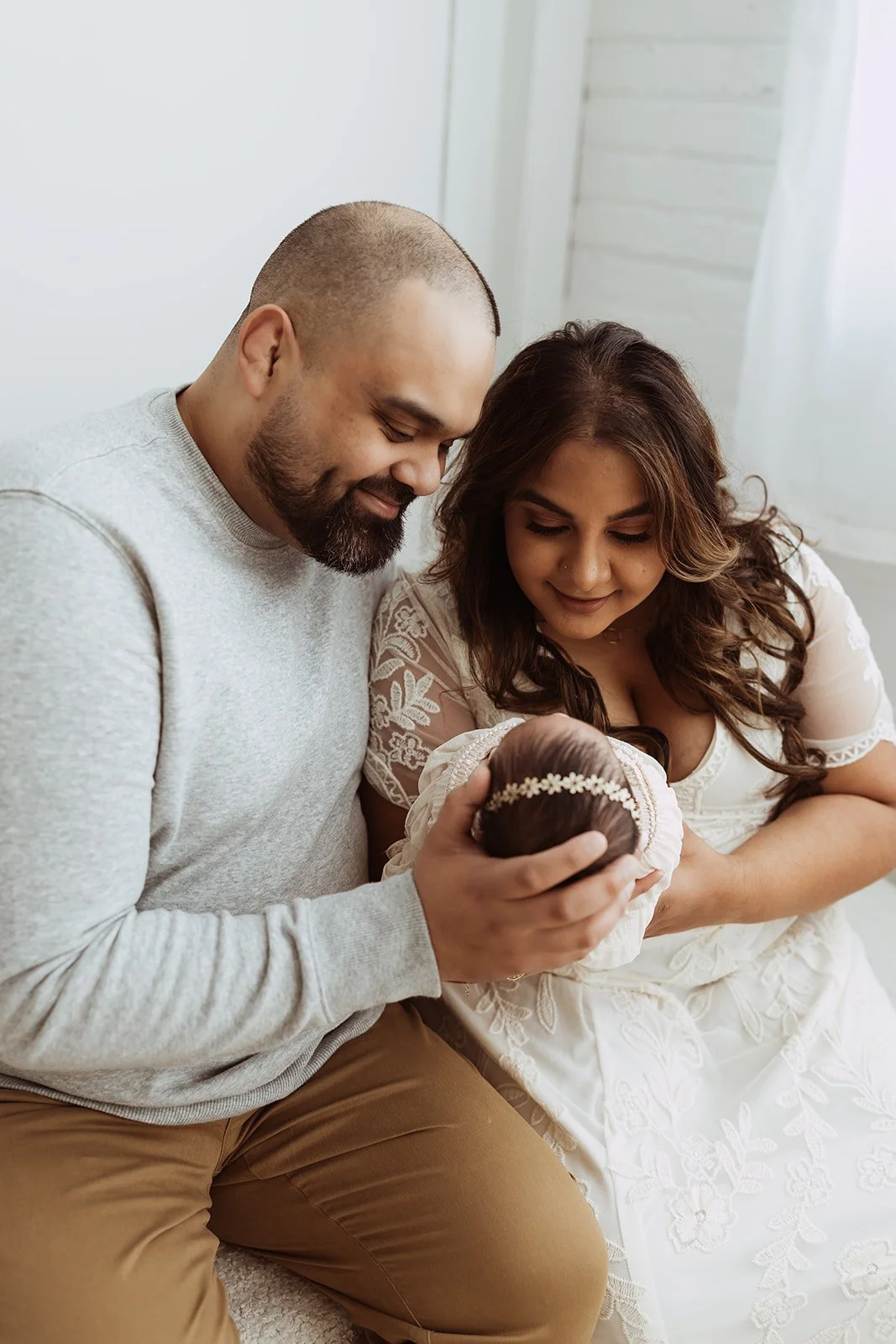  A mother and father hold their newborn baby girl in their laps, looking down at her lovingly. 