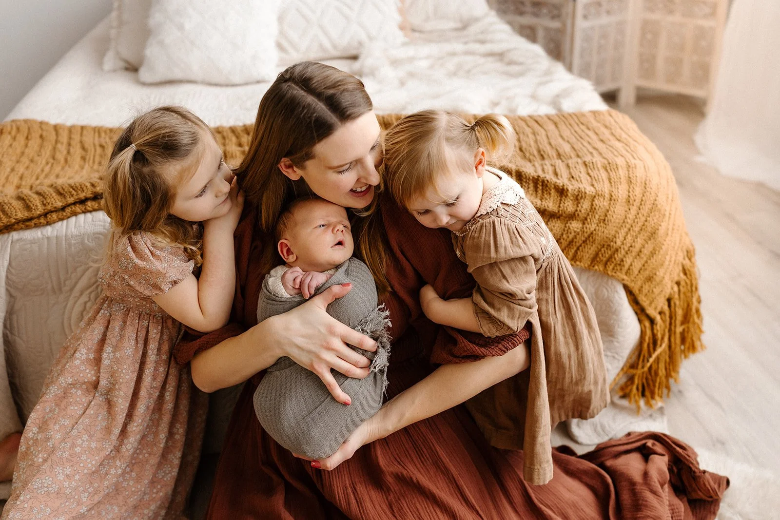  A mom holds her baby boy at the end of a bed. Her two toddler daughters surround her, giving her hugs. 