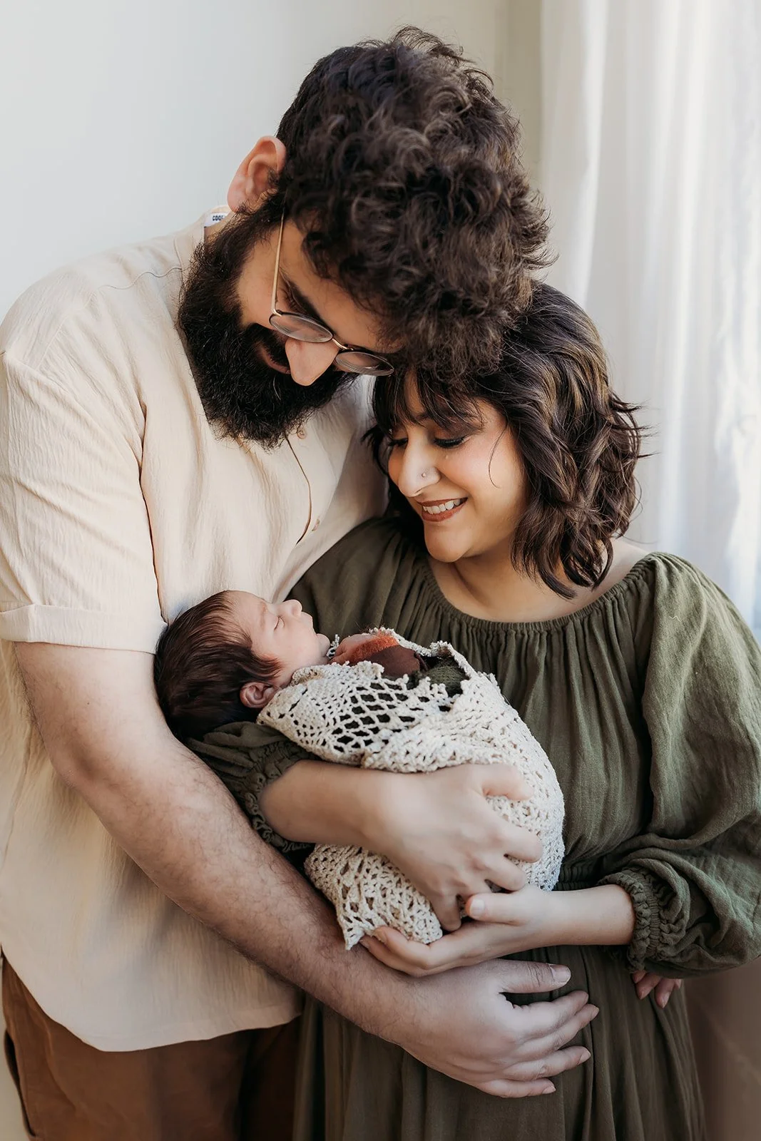  A mom and dad embrace and gaze at their newborn baby son, who they are holding between them. 