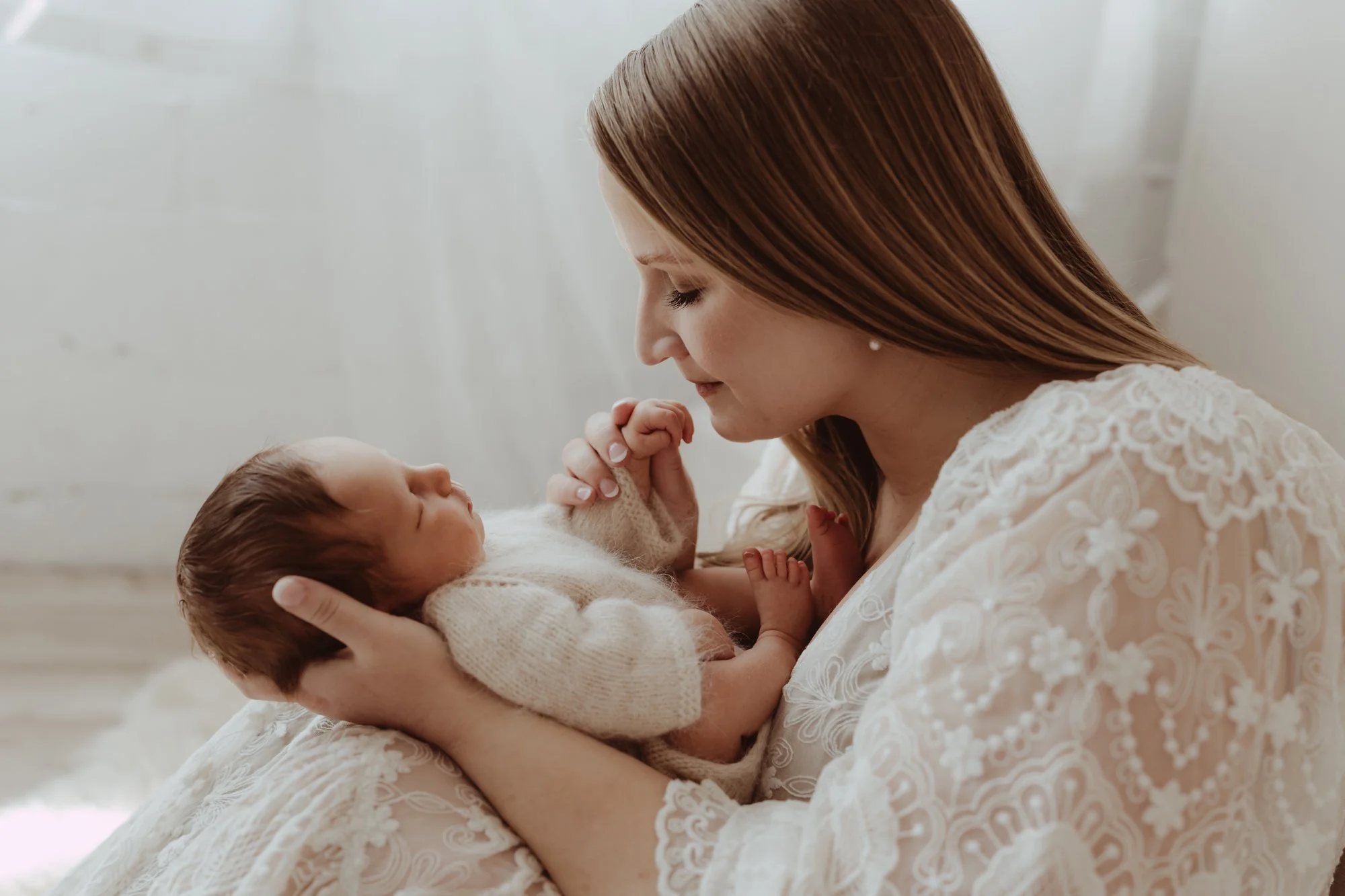 a mom in a white lace dress is kissing her baby girl's fingers during a newborn photography session in Minneapolis with AMG Photography