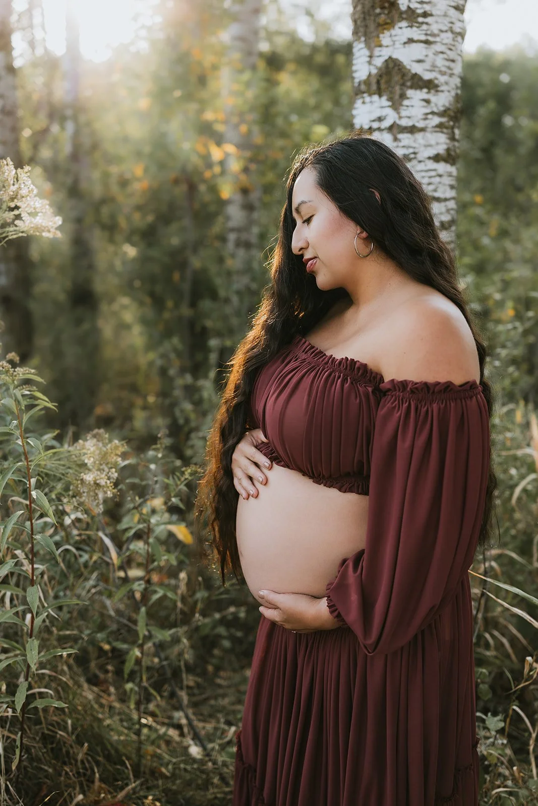 Pregnant woman in a burgundy off-shoulder dress standing outdoors in a forested area with sunlight filtering through the trees.
