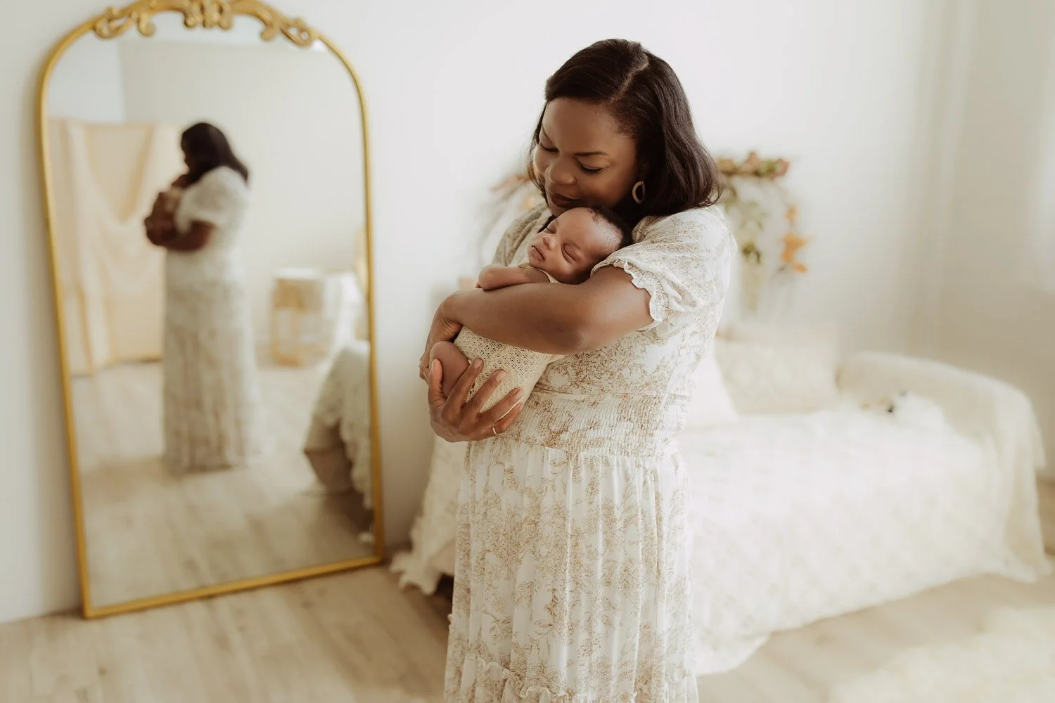 A mother holds her newborn baby girl in front of an ornate gold mirror during a lifestyle photo shoot.