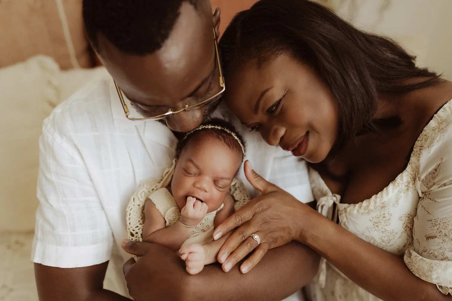 A mother and father sit on a bed and hold their newborn baby girl between them.