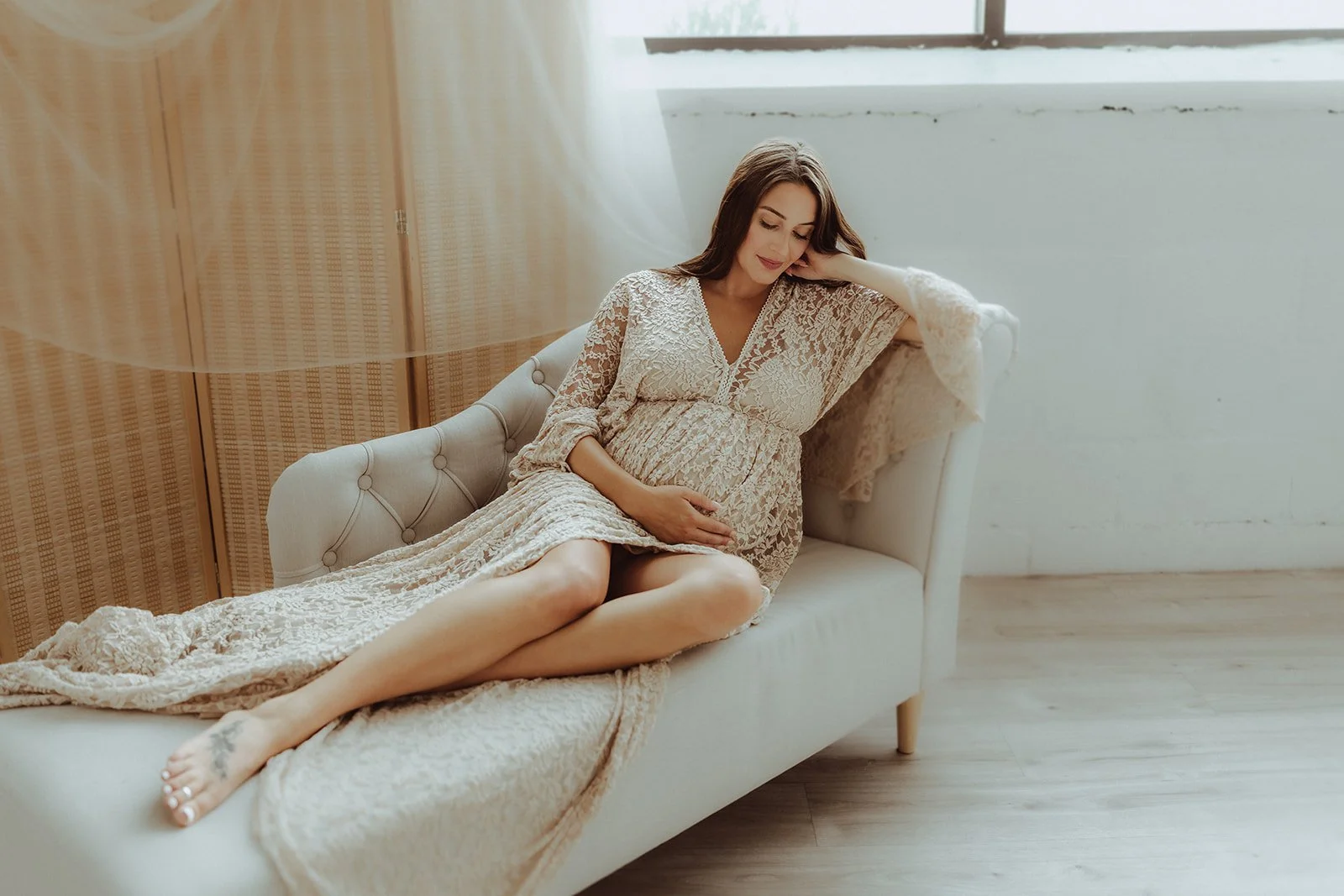A pregnant woman in a lacy dress reclining on a cream-colored sofa in a bright room.