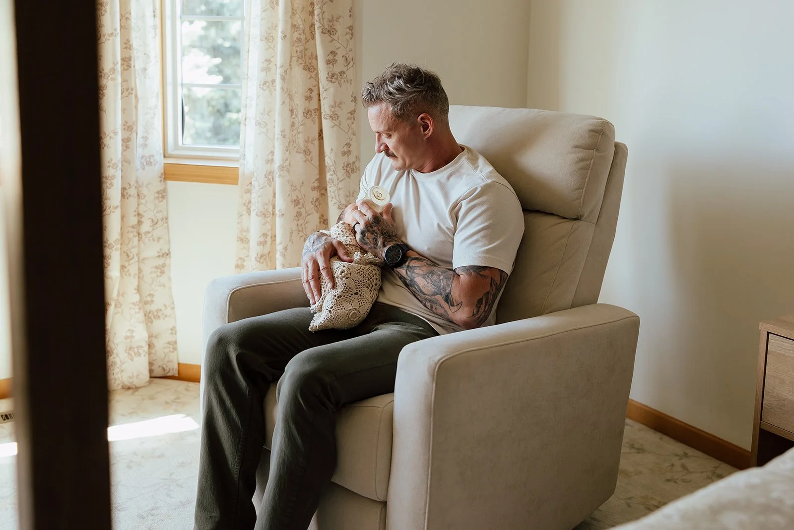 A man with tattoos on his arm sitting in a beige armchair holding a small dog wrapped in a crochet blanket, near a window with floral curtains.