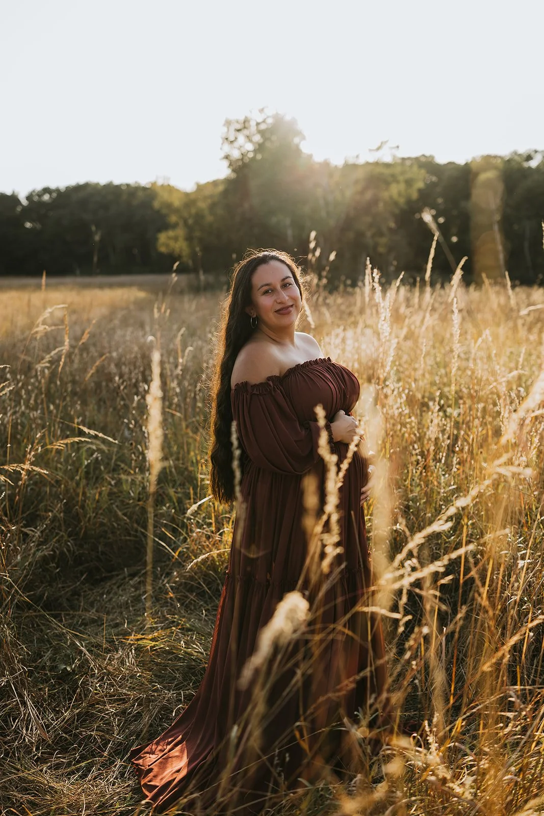 A woman with long dark hair wearing a brown off-shoulder dress stands in a field of tall, dry grass during sunset, smiling at the camera.