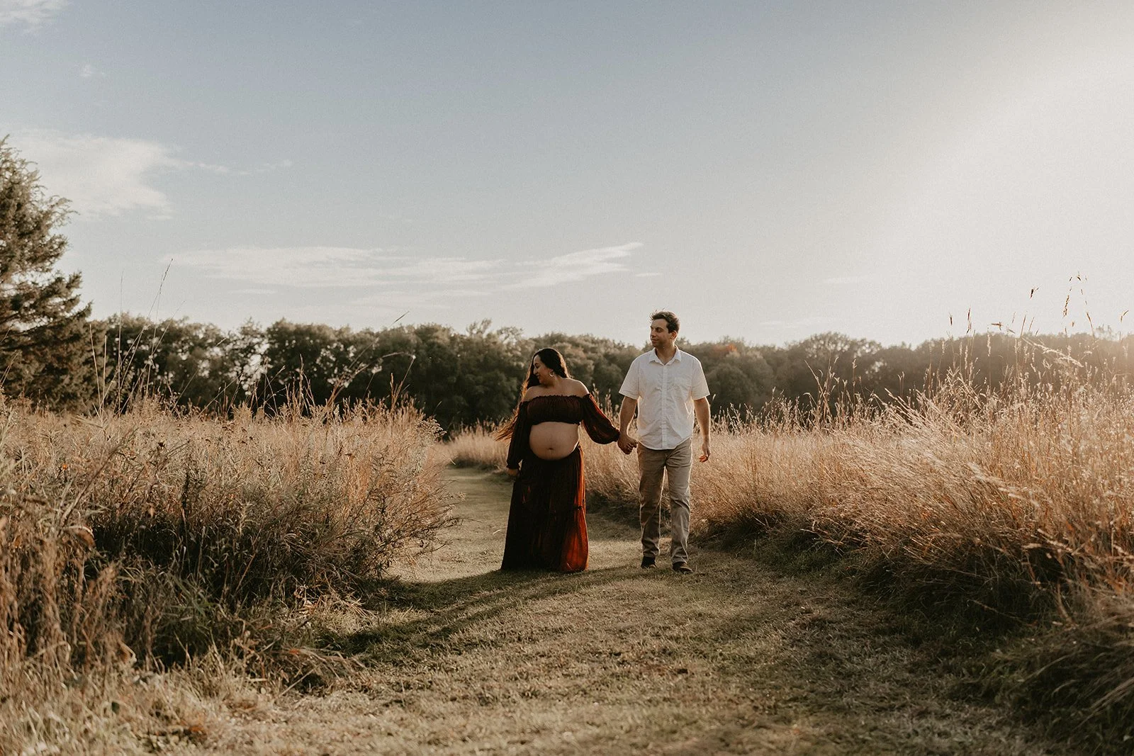 A pregnant woman and a man holding hands walk on a grassy path through a field of tall, golden grass with trees in the background during sunset.
