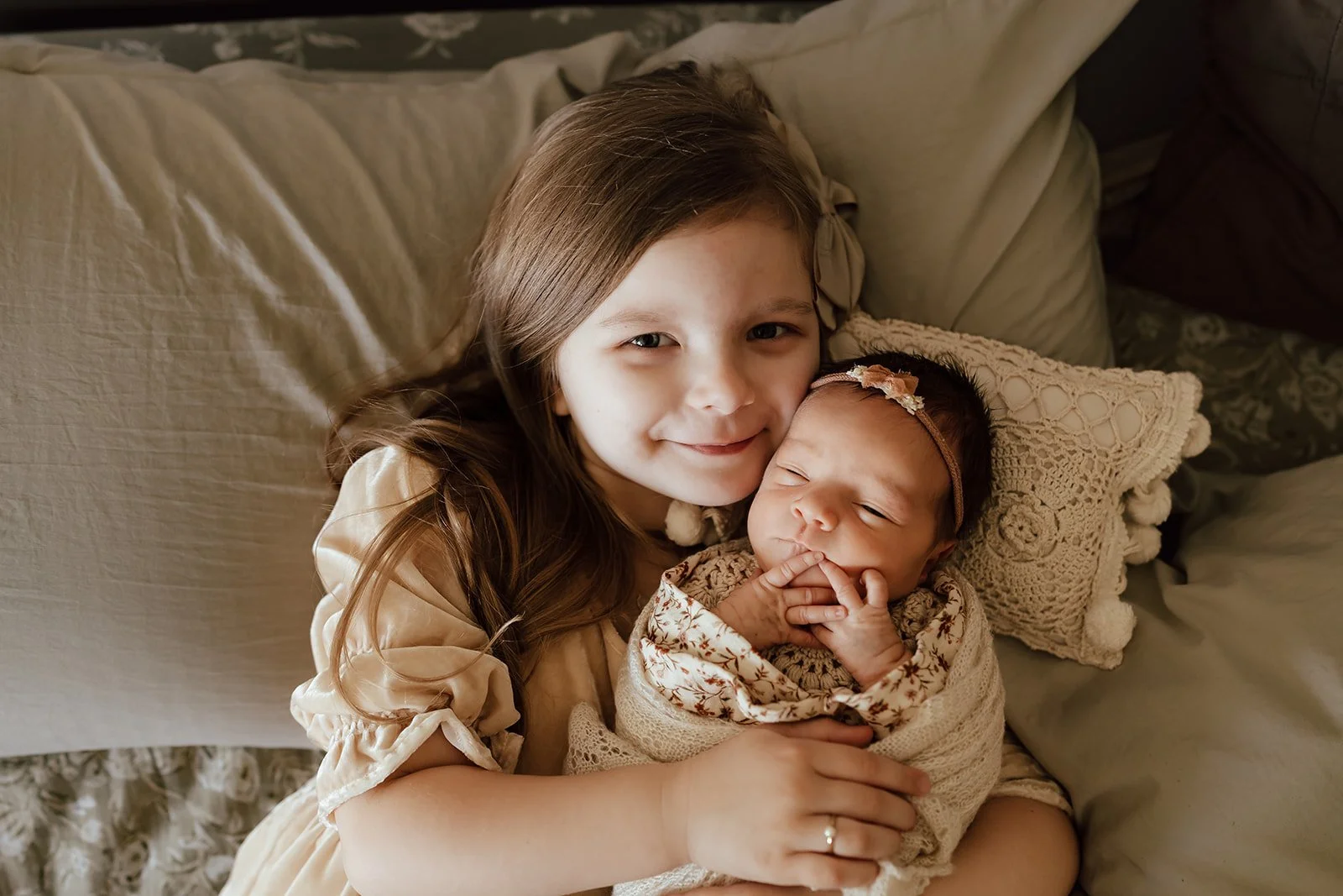A young girl smiling and holding a newborn baby girl with a pink headband, sitting on a bed with pillows.