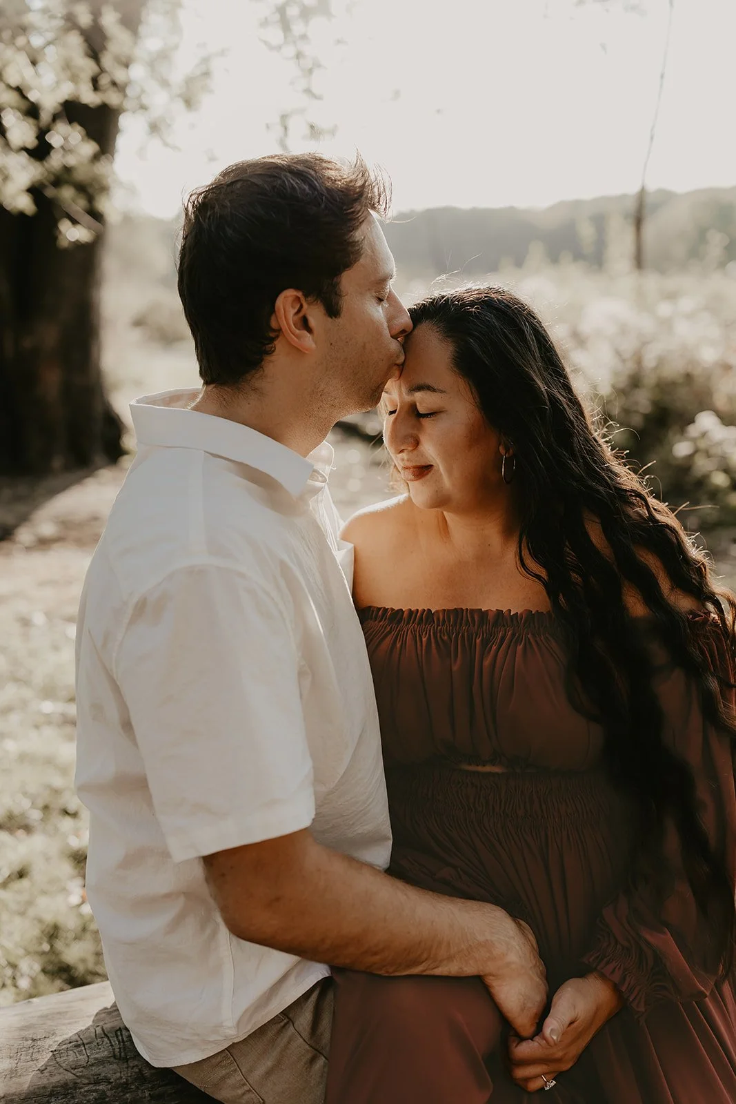 A man kissing a woman on the forehead outdoors, with natural sunlight and blurred trees in the background.