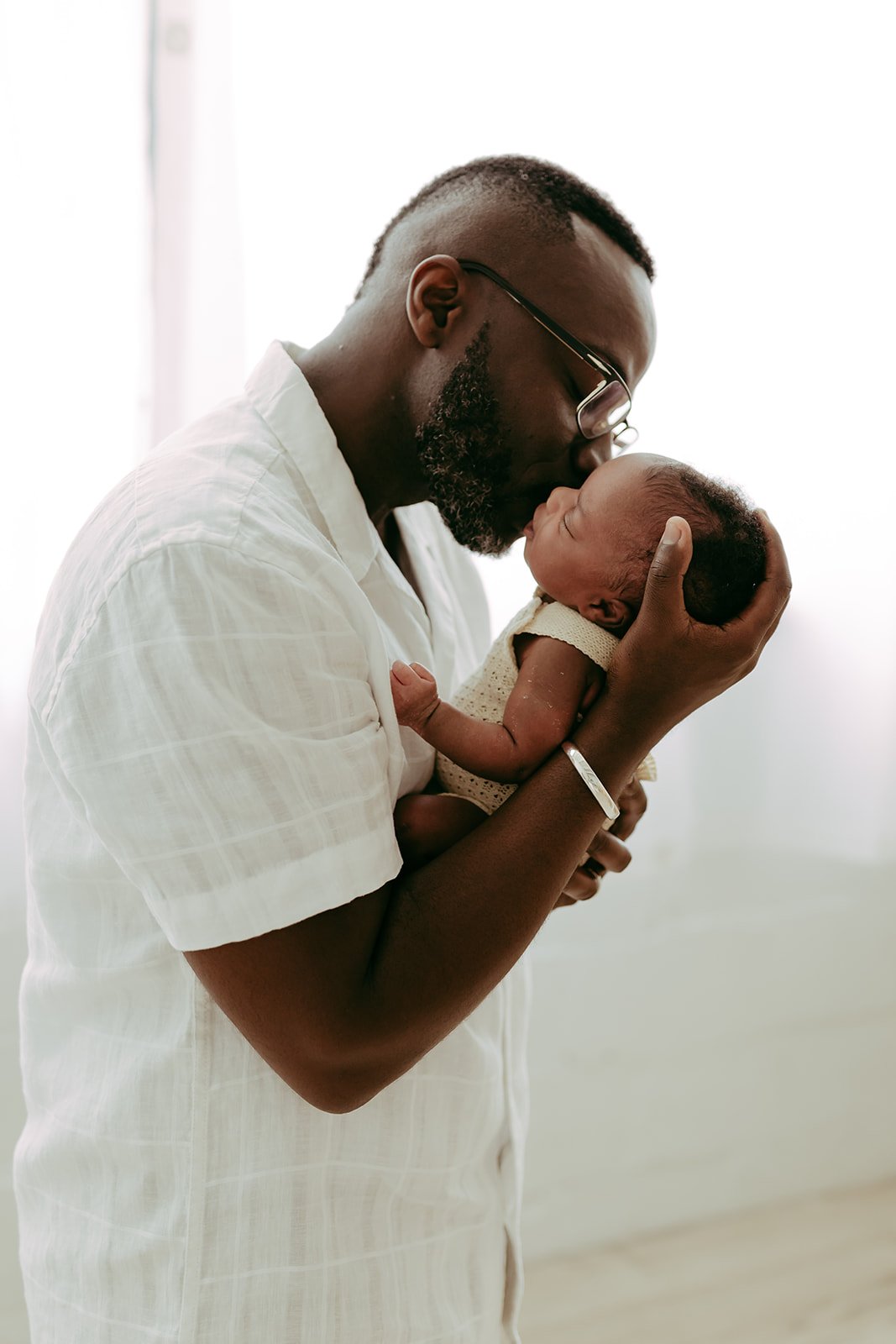 A dad holds his newborn daughter up and kisses her cheek in front of a window with white curtains.