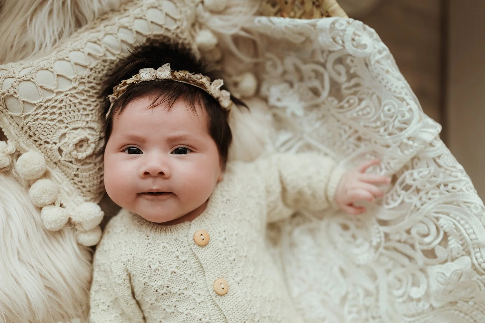 Adorable baby girl with dark hair, wearing a cream-colored knitted cardigan with wooden buttons and a delicate floral headband, lying on a cozy, cream-colored blanket with intricate lace patterns.