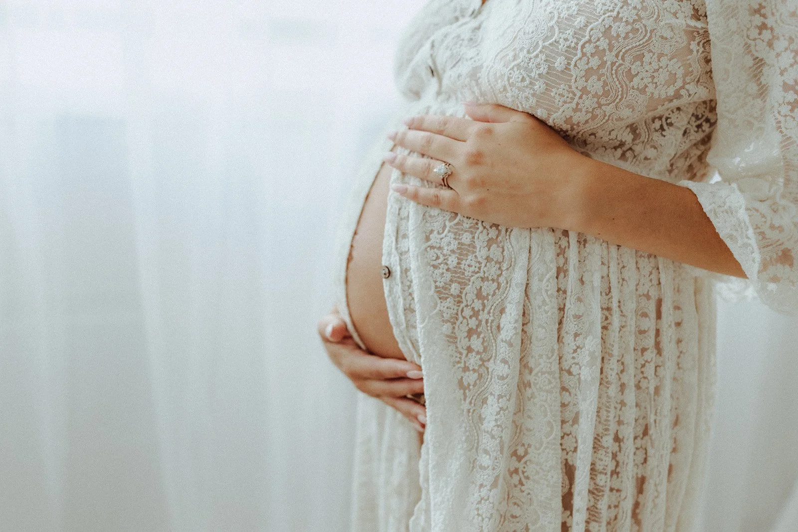 A pregnant woman wearing a lace dress, gently holding her belly with one hand and showing an engagement ring on her finger, in a softly lit setting.