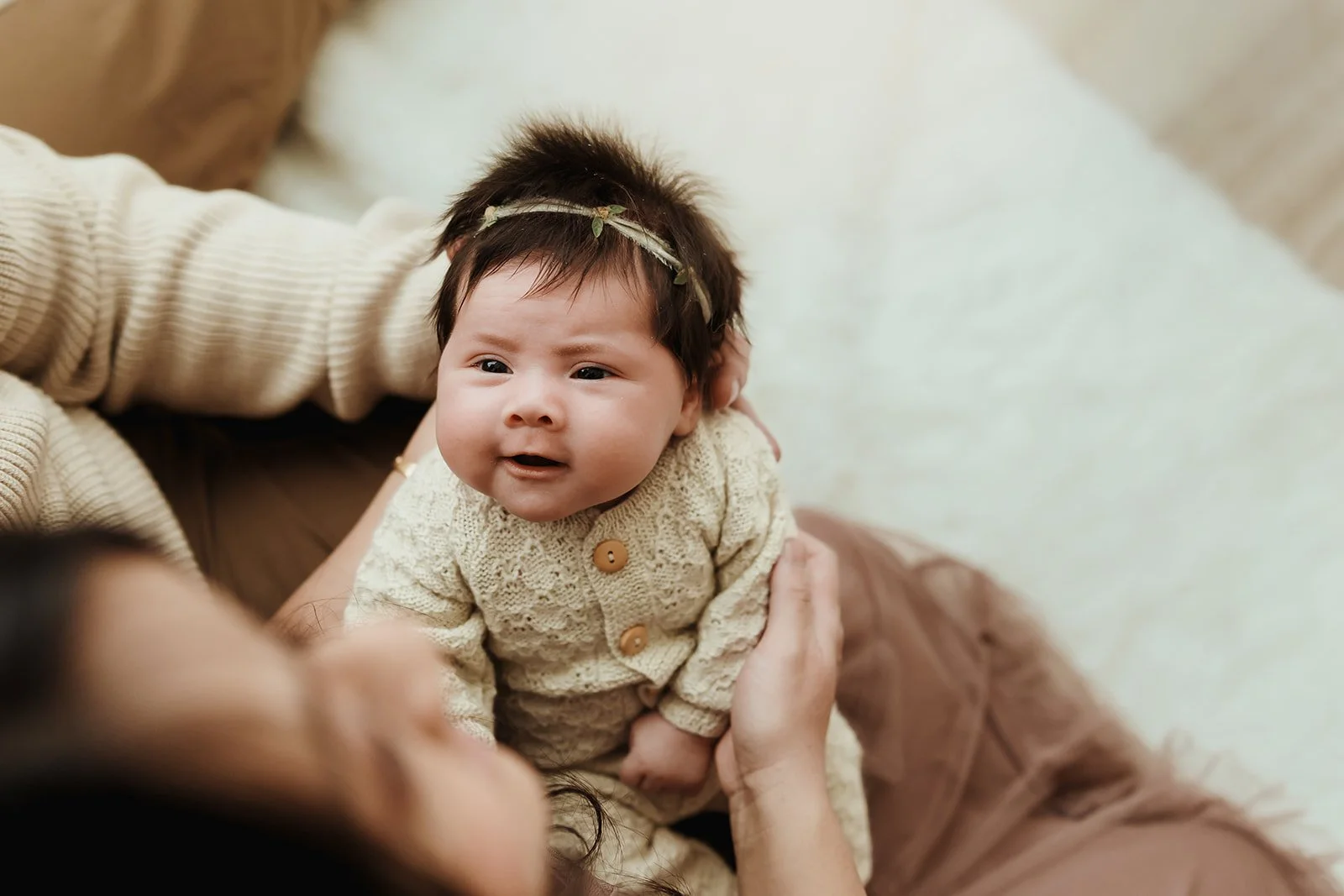 A baby girl with brown hair and a headband, lying on an adult's chest, looking up with a curious expression.