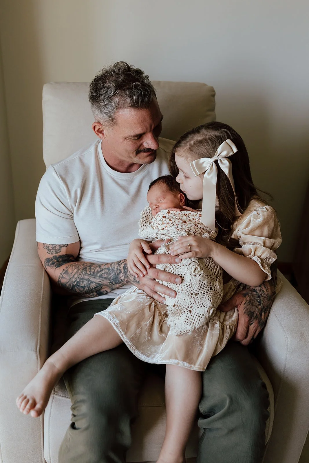A man with tattoos holding a young girl and a newborn baby in a cozy room.