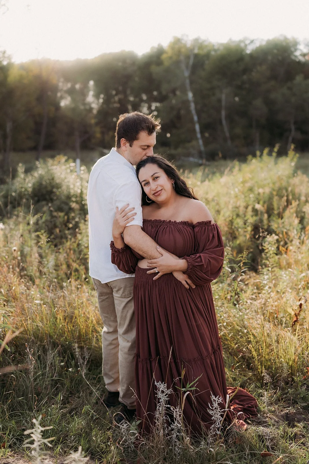 A couple stands close together in a grassy field during sunset, with trees in the background. The woman is wearing a long, off-the-shoulder maroon dress, and the man is dressed in a white shirt and beige pants. They are embracing affectionately, look