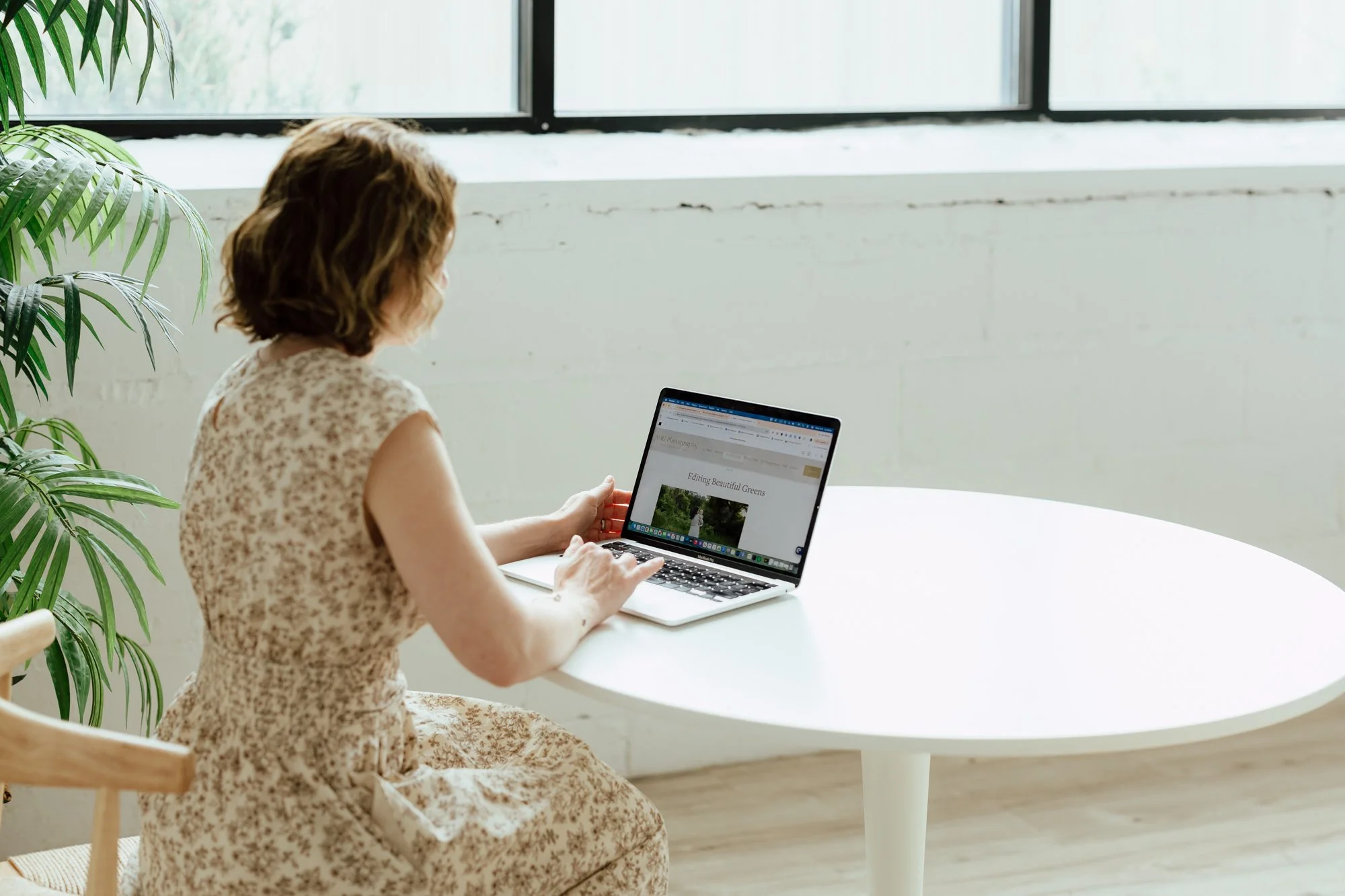 A woman with curly brown hair wearing a beige floral dress, sitting at a white table with a laptop, in a bright room with white walls and large windows, next to a green leafy plant.