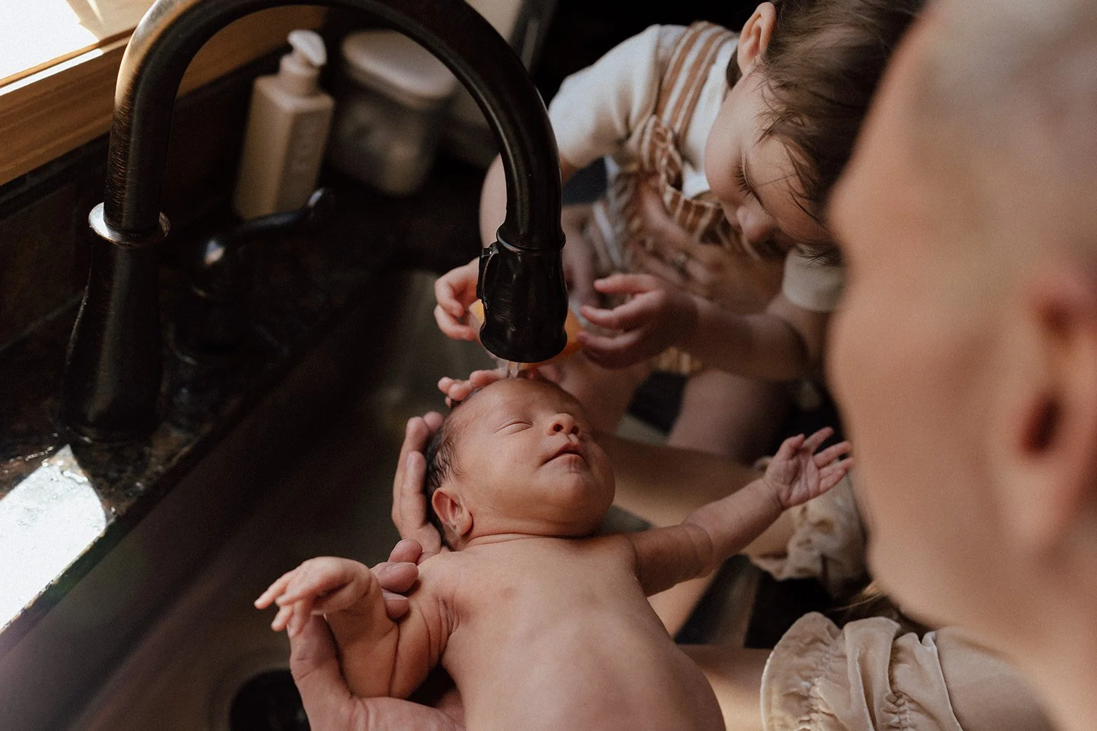 A woman is bathing a newborn baby in a kitchen sink. Two children are nearby, watching and leaning in to observe the bathing process.