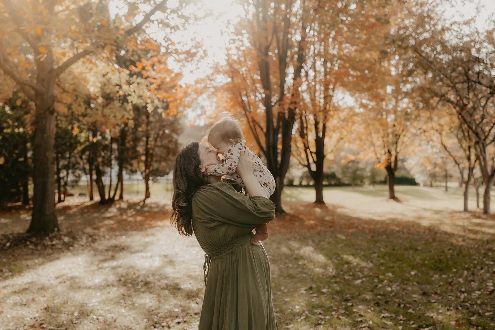 A woman holding a young child in a park during autumn with trees showing orange and yellow leaves