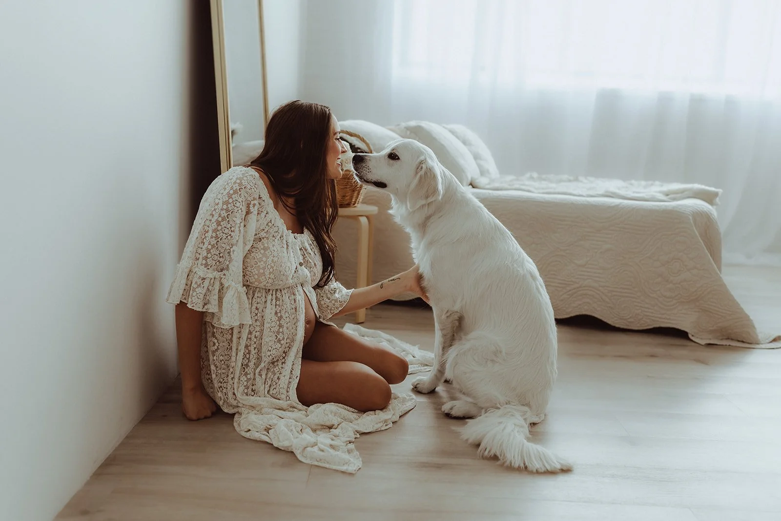 A woman with long brown hair, dressed in a cream lace dress, sits on the floor of a bedroom, facing a large white dog. They are touching noses affectionately, with the woman smiling. The room has light-colored wooden flooring, a bed with a textured c