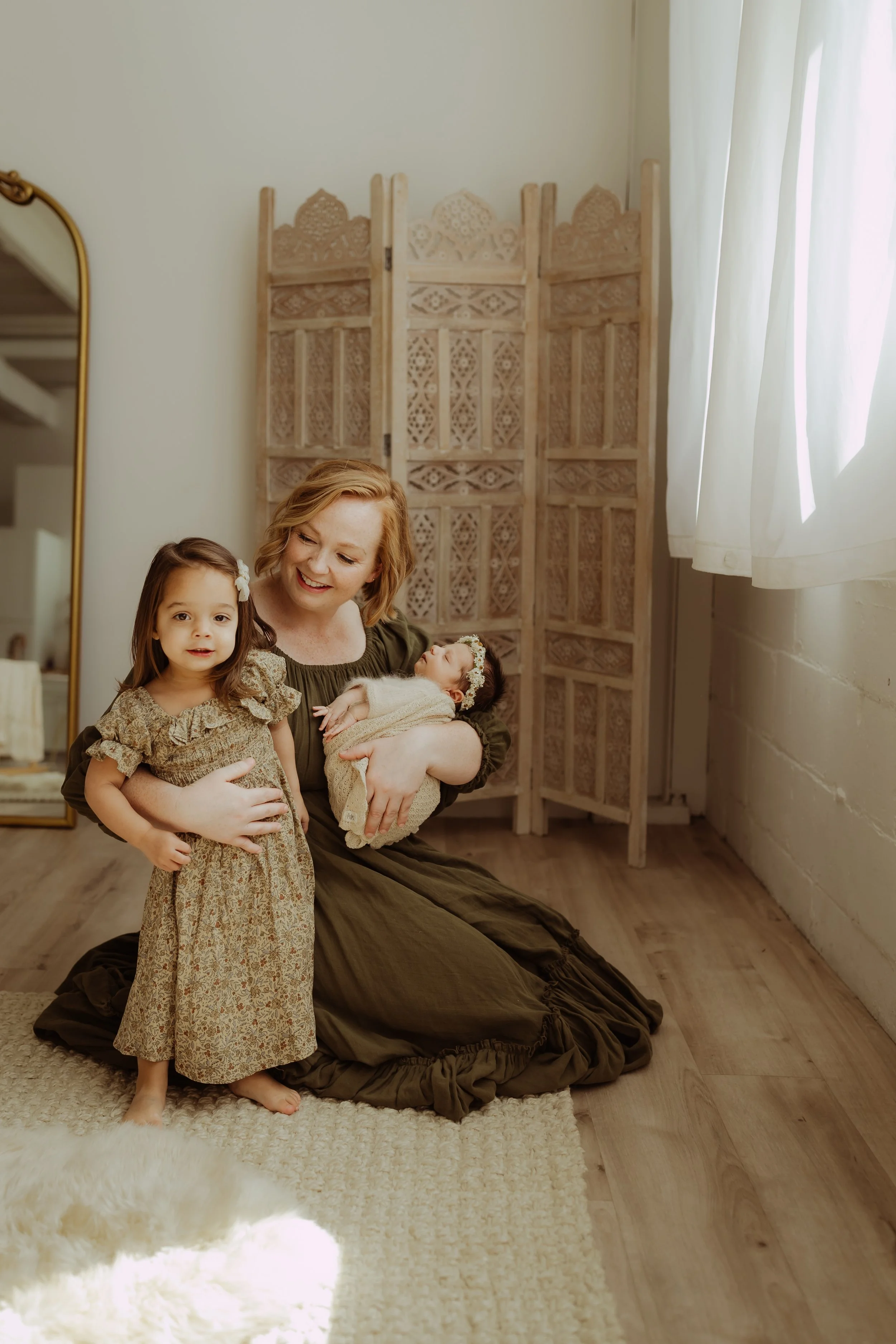 A woman with short hair kneeling on a wooden floor indoors, wearing a green dress, smiling, and holding a baby wrapped in a blanket. Next to her is a young girl in a floral dress. There is a decorative wooden screen and a rug in the background.