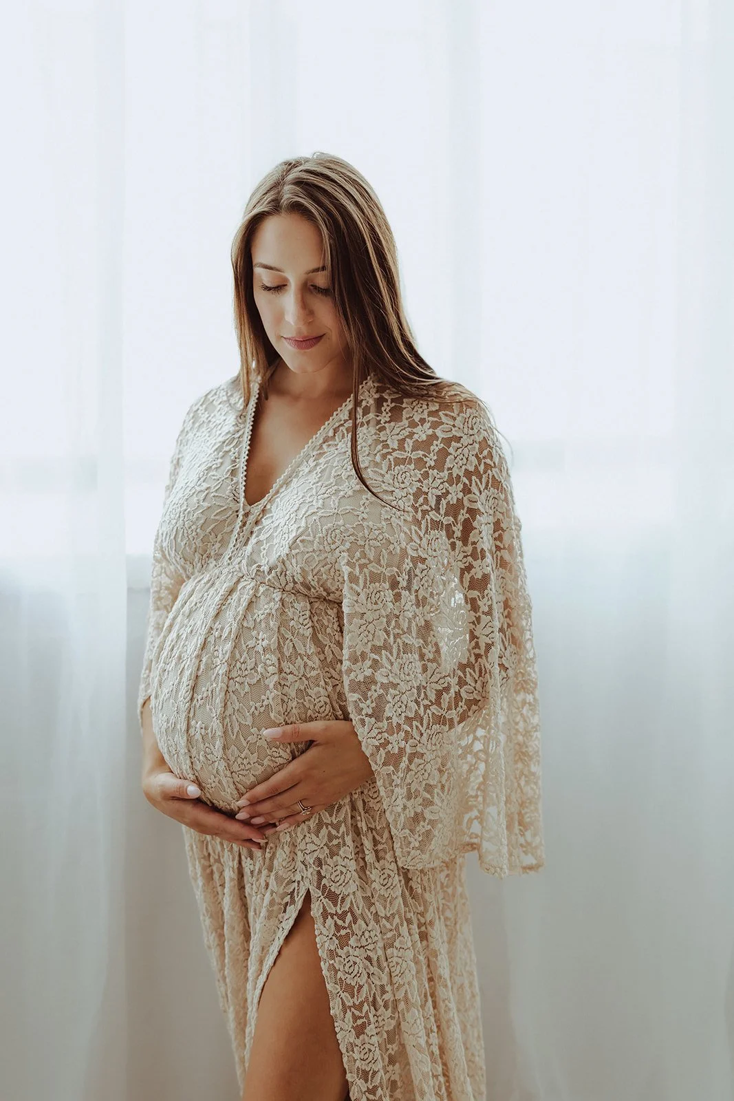 A pregnant woman with long brown hair, standing by a window with sheer curtains, wearing a beige lace robe and holding her belly with both hands, looking down peacefully.