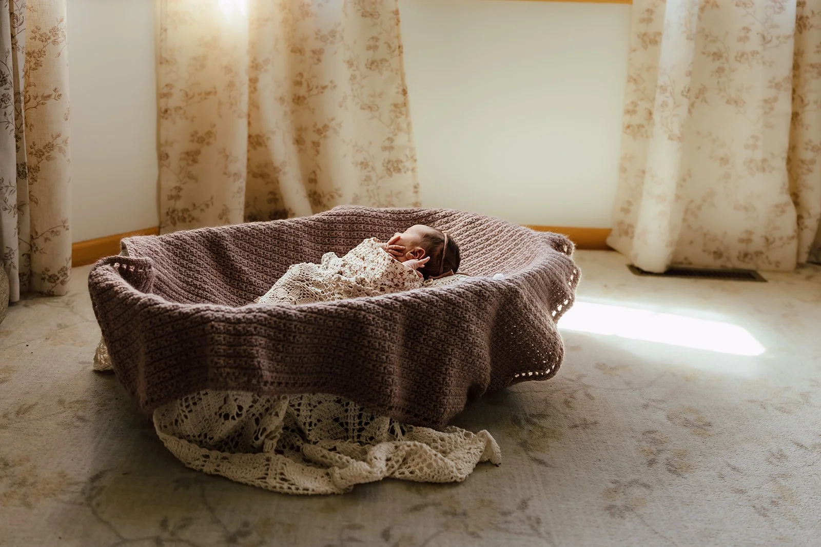 A young child or baby sleeping in a cozy, knitted crib with a blanket, in a room with floral curtains and a light-colored carpet.