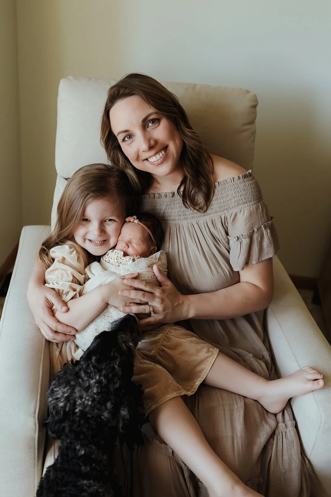 A woman sitting on a beige armchair with three young girls and a black puppy, all smiling, in a room with beige walls.