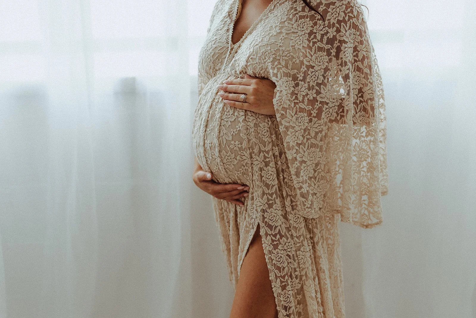 Pregnant woman in a beige lace dress standing against a light background, cradling her belly with one hand, showing an engagement ring on her finger.