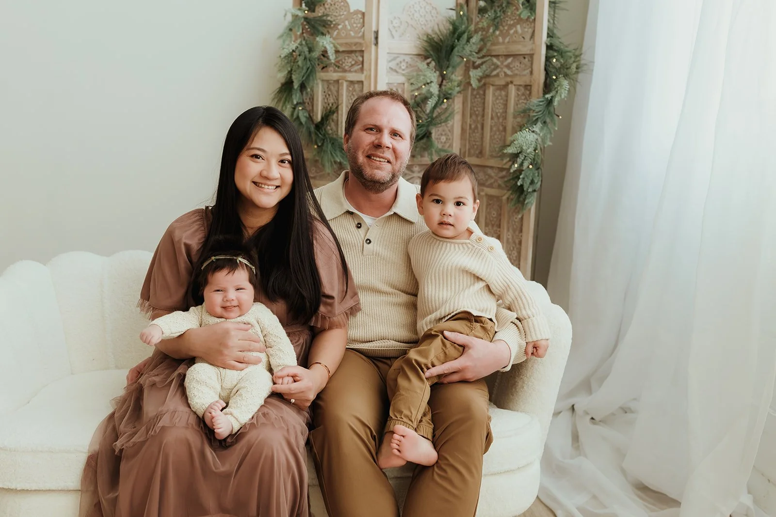 A family of four sitting on a cream-colored couch in a decorated living room. The woman on the left, with long black hair, is smiling and holding a smiling baby girl dressed in cream. The man in the middle, with short hair and a beard, is smiling and