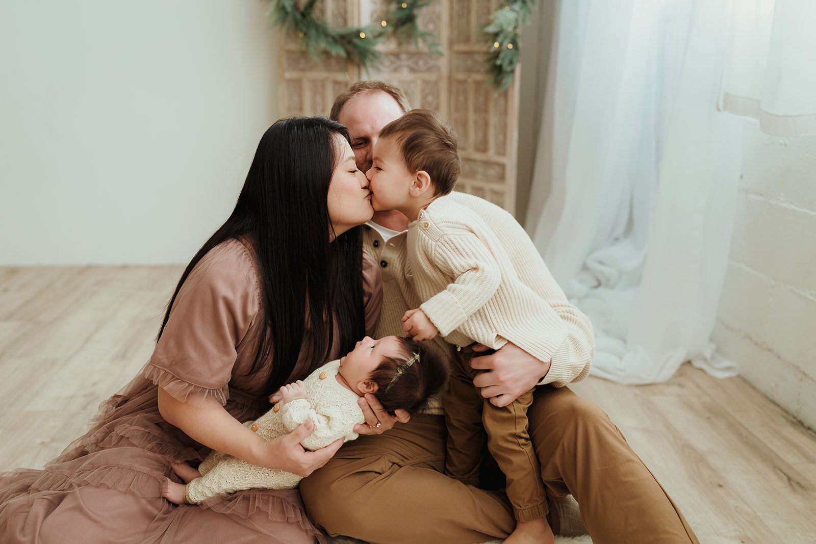Family of four, including a woman, a man, a young boy, and a baby girl, sharing a kiss in a cozy, decorated room with natural light.