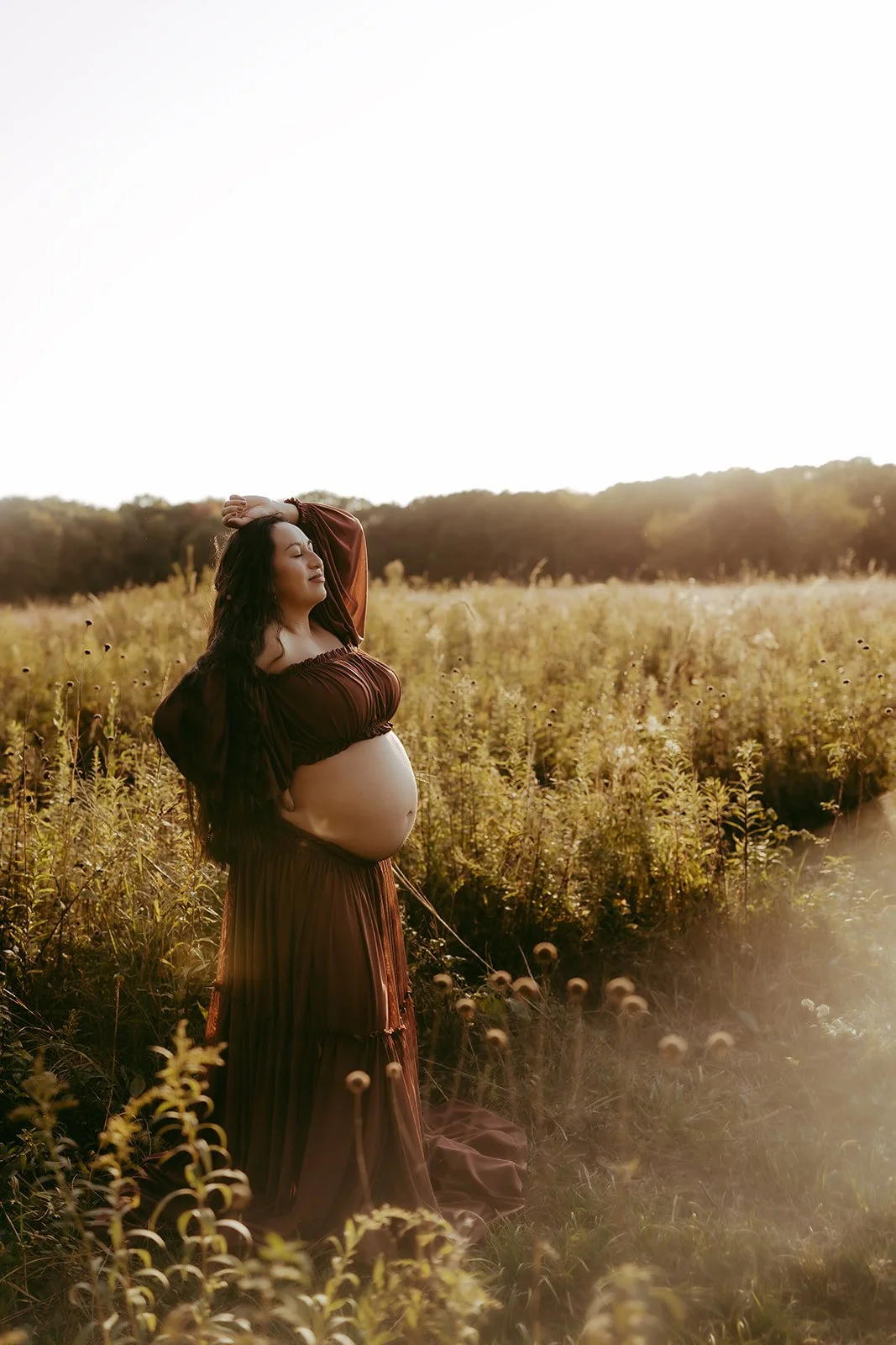 Pregnant woman in a brown dress standing in a grassy field during sunset, with her eyes closed and arms raised over her head.