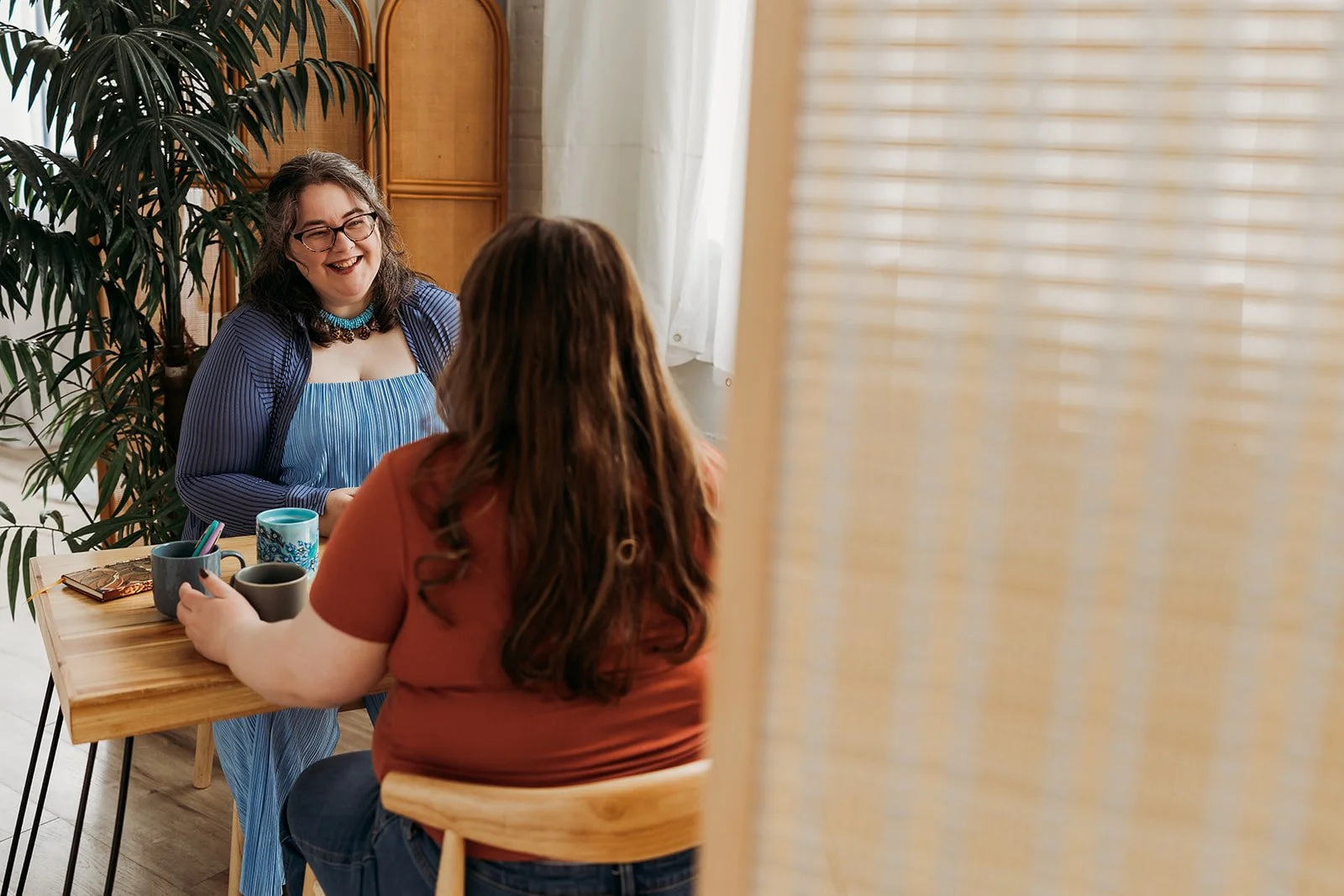 Two women sitting at a table having a conversation in a cozy room with plant decor. One woman is smiling, wearing glasses, a blue-striped dress, and a blue cardigan. The other has long hair, wearing a reddish-orange top. The table holds coffee mugs and a notebook.