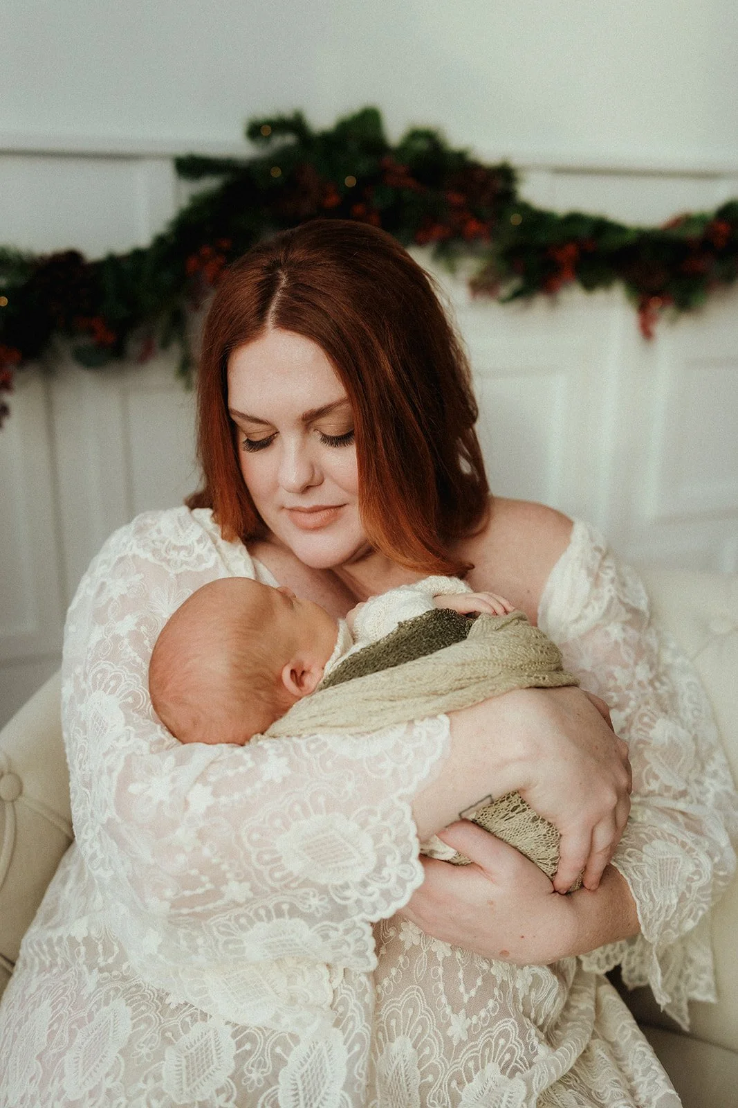 A woman with red hair in a white lace dress holding a sleeping baby wrapped in a green and cream blanket. They are sitting on a white couch with a holiday garland behind them.
