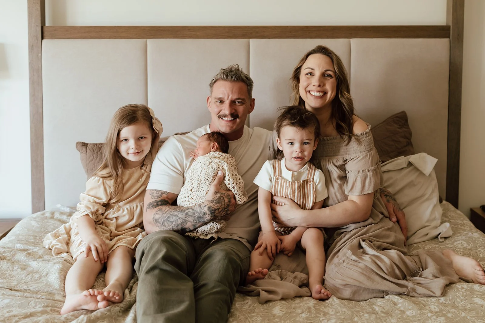 A smiling family of six sitting on a bed in a cozy bedroom. The family includes two adults, a man holding a newborn baby, a woman, and three young girls. The man has tattoos and the woman has long wavy hair. The children have different hair colors an
