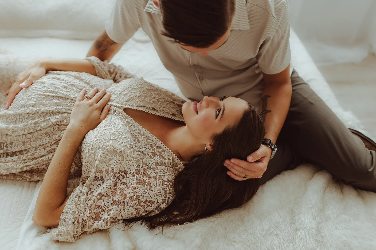 A woman lying on a bed with her head resting on a man’s lap, gazing up at him; the man leaning over her, holding her head gently, in a cozy, intimate setting with soft lighting.