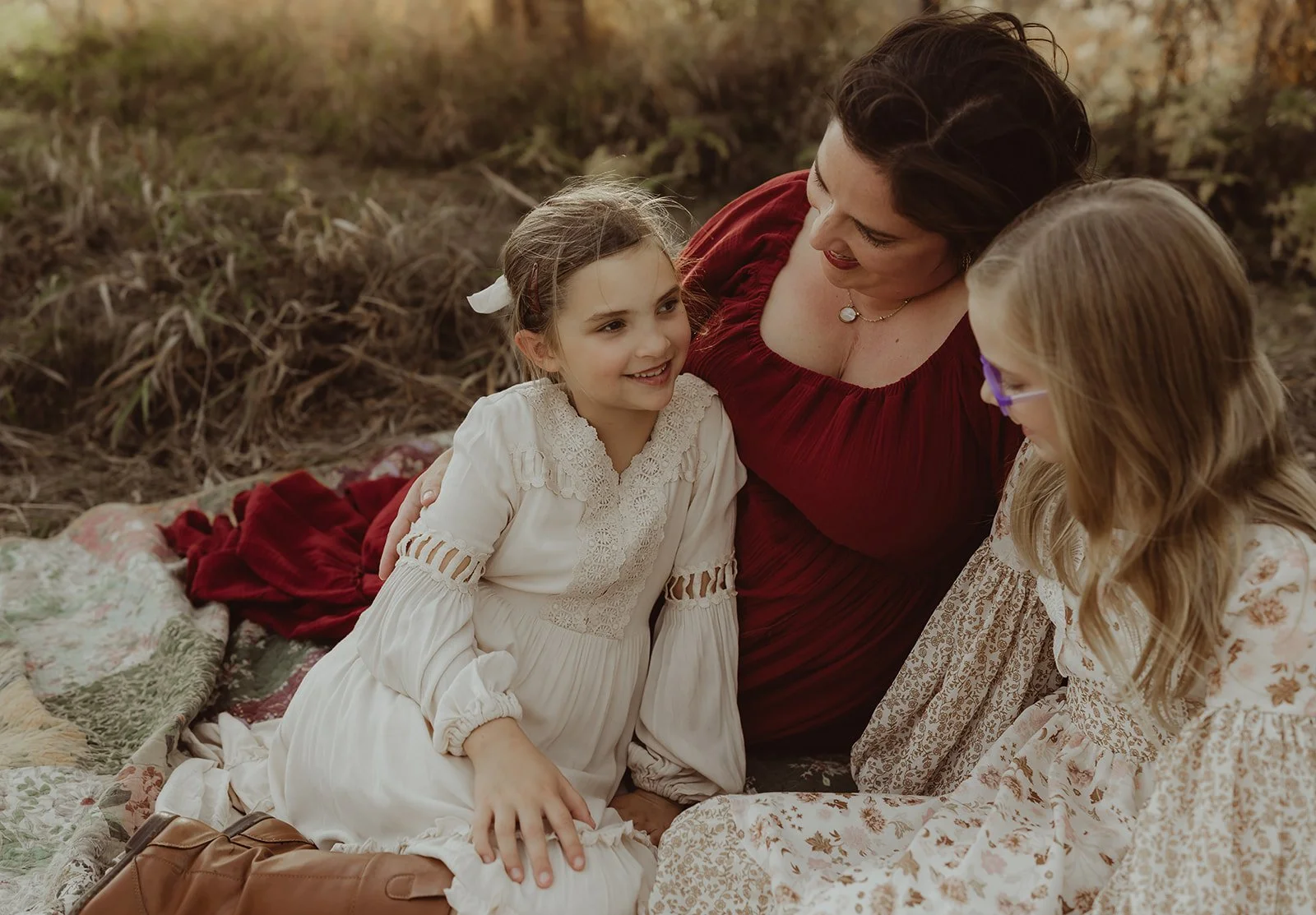Three women sitting on a blanket outdoors, smiling and talking, with trees and foliage in the background.